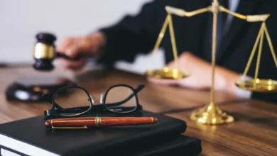 Önünde siyah defter, gözlük ve kalem bulunan masada adalet terazisi ile hakim tokmağı tutan kişi.