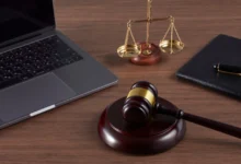 Ahşap masa üzerinde dizüstü bilgisayar, hakim tokmağı, adalet terazisi ve siyah defter.