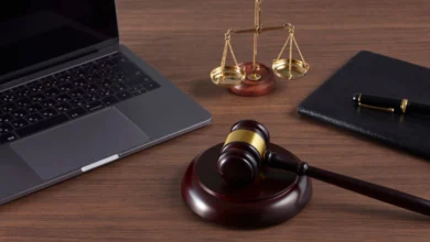 Ahşap masa üzerinde dizüstü bilgisayar, hakim tokmağı, adalet terazisi ve siyah defter.